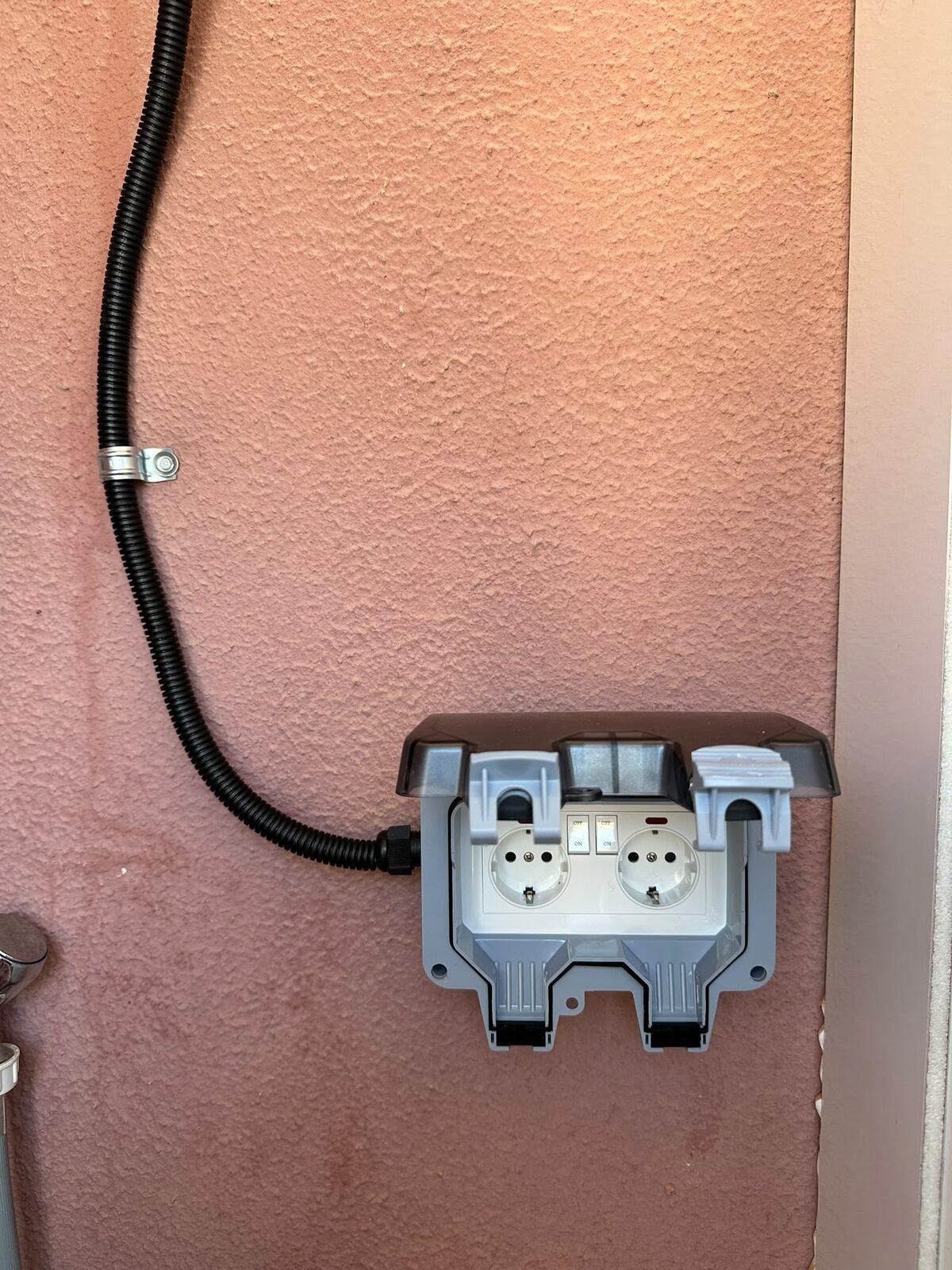 Base d'endoll doble estanca amb tapa instal·lada en una façana exterior, alimentada amb tub corrugat negre fixat amb abraçadora metàl·lica